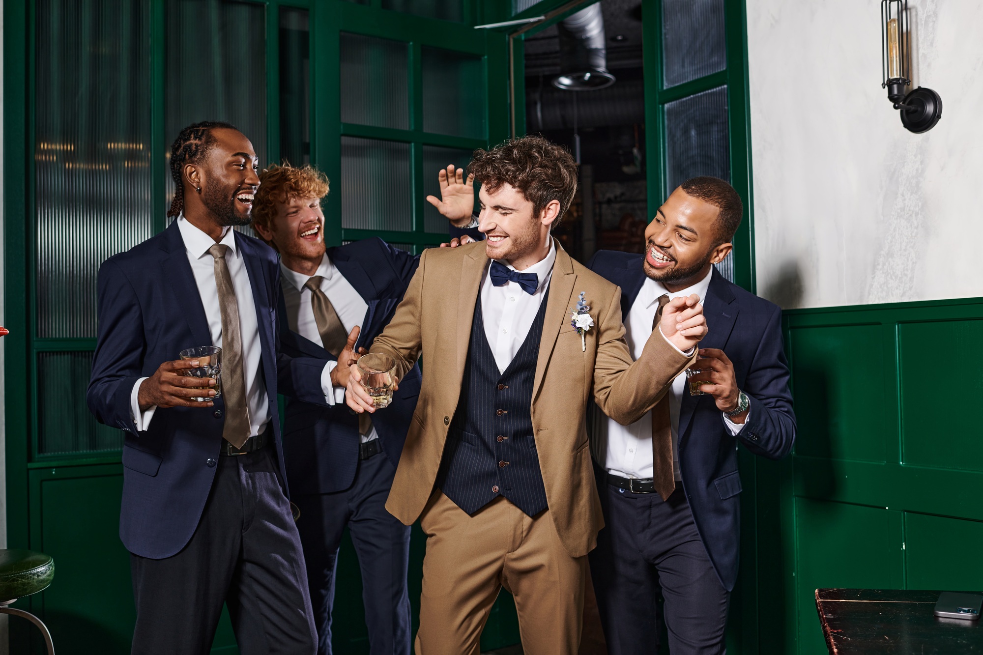 bachelor party, multiethnic best men and groom laughing while standing with glasses of whiskey